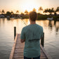 Man on a dock holding a fishing rod with a sunset and palm trees in the background, wearing a 'Dogfish Tackle & Marine' t-shirt.