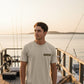 Man standing on a dock with fishing rods, wearing a t-shirt with a logo, during sunset.
