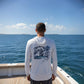 Person wearing a white long-sleeve shirt with a graphic design, standing on a boat with ocean and sky in the background.