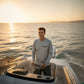 Man on a boat wearing a gray long-sleeve shirt with a logo, steering the boat at sunset.