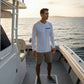 Man standing on a boat wearing a white hoodie with a logo, looking out over water at sunset.