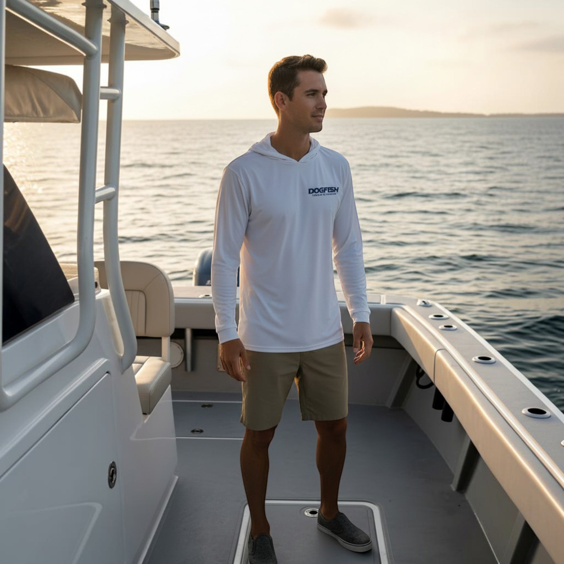 Man standing on a boat wearing a white hoodie with a logo, looking out over water at sunset.