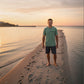 Man standing on a beach at sunset wearing a green t-shirt and dark shorts.