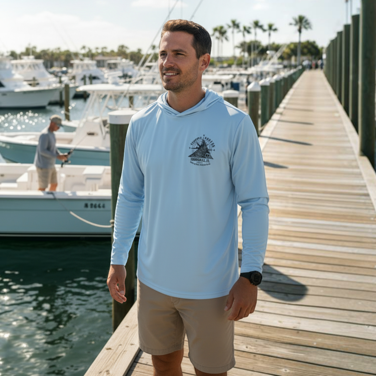 A man wearing a light blue Dogfish Tackle & Marine long sleeve hooded performance shirt on a sunny marina dock.