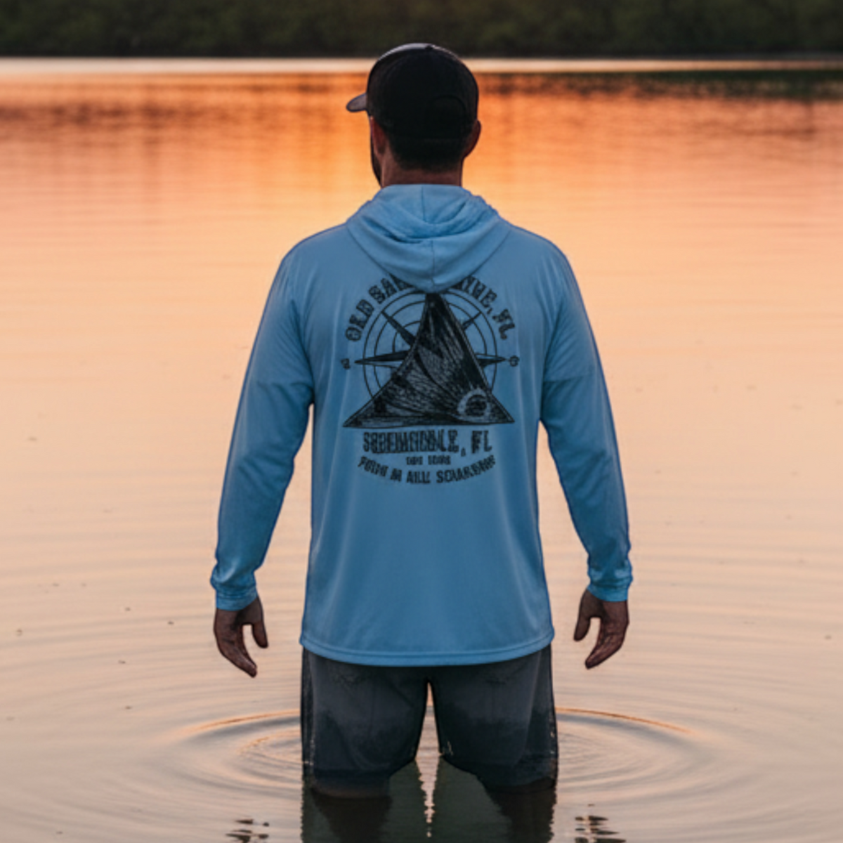 Man wearing a Dogfish Tackle & Marine light blue long sleeve performance hoodie standing in water at sunset.
