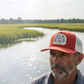 Dogfish Tackle & Marine Marsh Wear orange and white trucker hat with woven patch worn by a man near a marsh