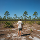 Man wearing a cream Marsh Wear Mission T-Shirt with a back graphic, sold by Dogfish Tackle & Marine in a pine forest.