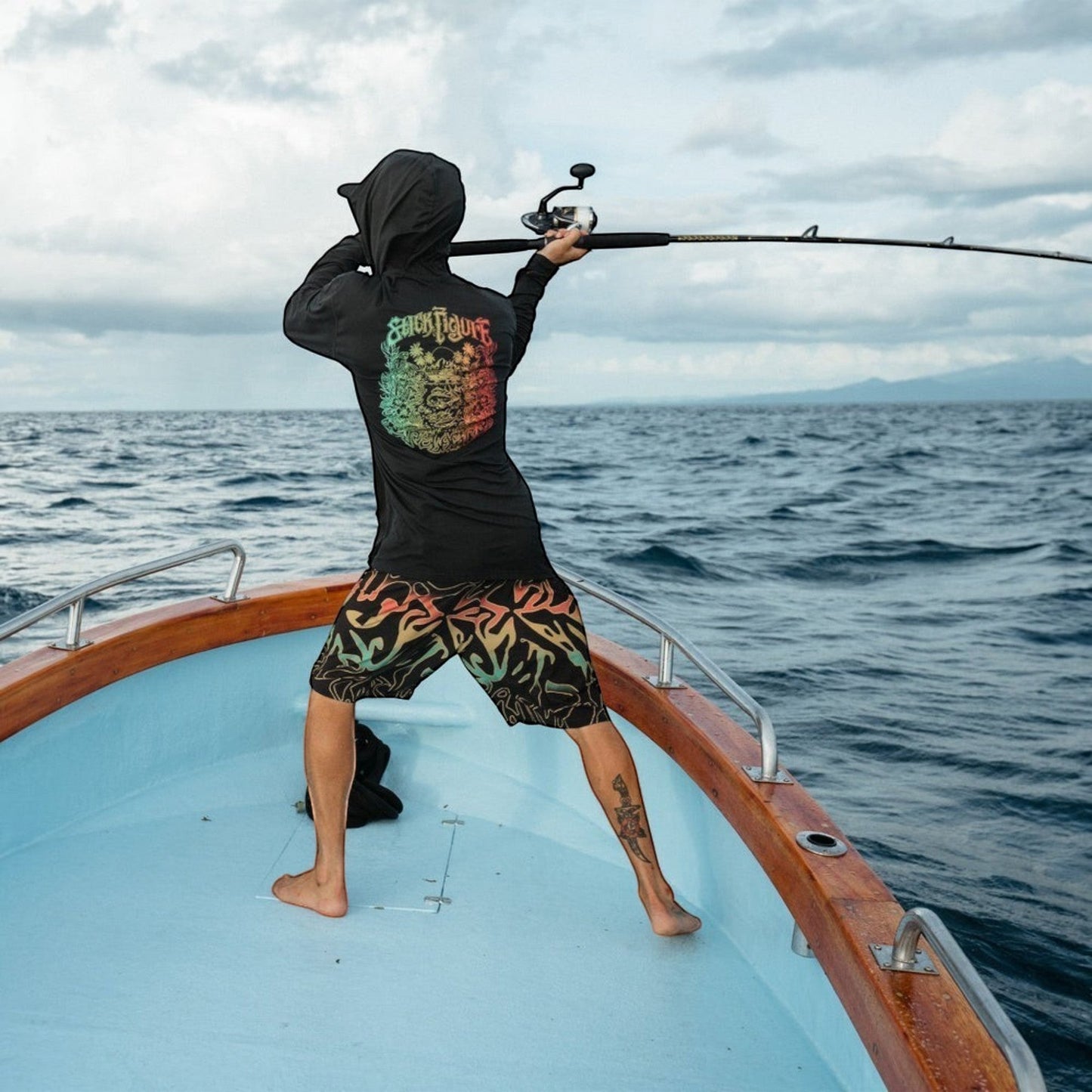 Dogfish Tackle & Marine man wearing black boardshorts with colorful wavy pattern, fishing on a boat.