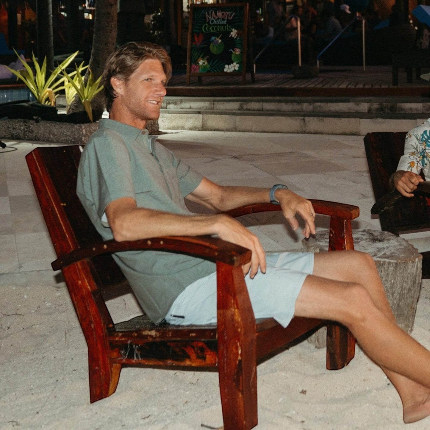 Dogfish Tackle & Marine man in a sage green short-sleeve button-down shirt and light blue shorts sitting in a wooden chair on the beach.