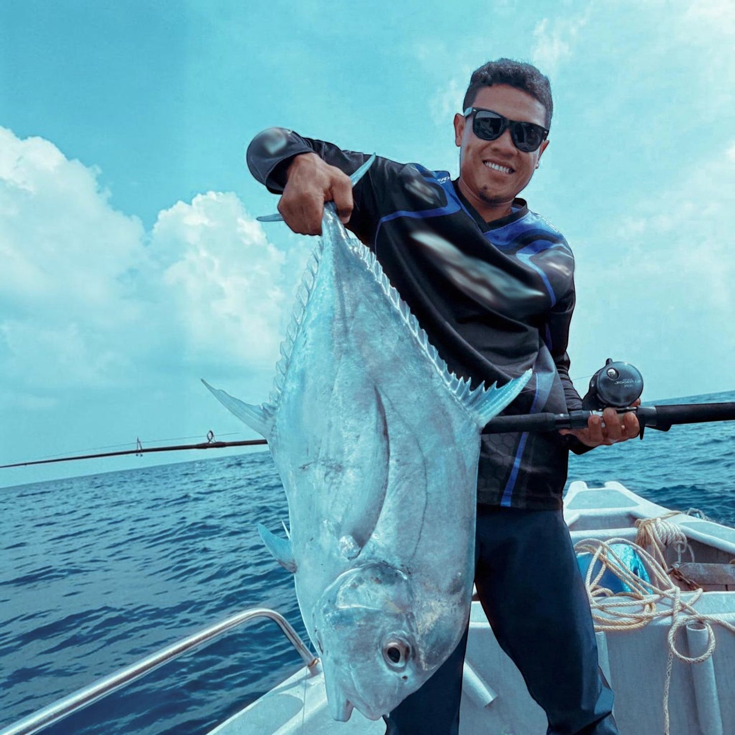 Dogfish Tackle & Marine angler holding a large silver fish on a boat, blue sky with clouds.