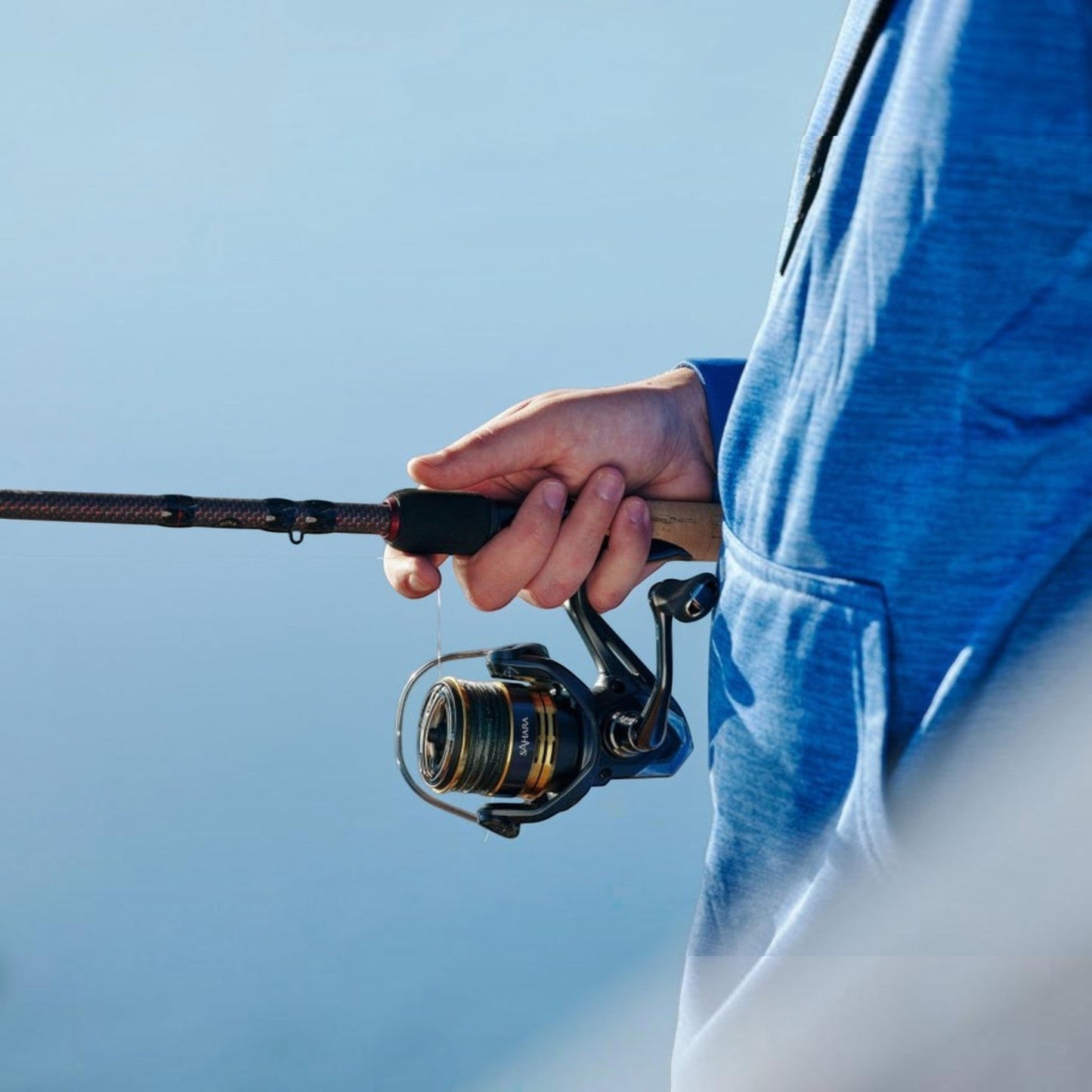 Dogfish Tackle & Marine Shimano Sahara FJ spinning reel with gold and black spool, held by a person in a blue hoodie.